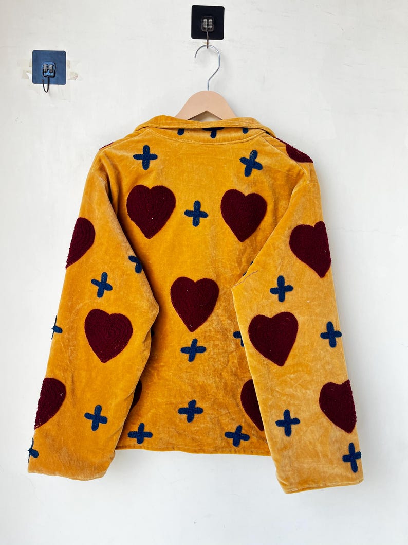 May include: Mustard yellow jacket with a velvet texture, featuring large burgundy heart appliqu&eacute;s and navy blue cross accents. The jacket is hanging on a wooden hanger against a white wall.