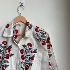 May include: Cream-colored pajama top with a red floral pattern and a white trim. The long-sleeved top has a button-down front, a chest pocket, and is hanging on a wooden hanger.