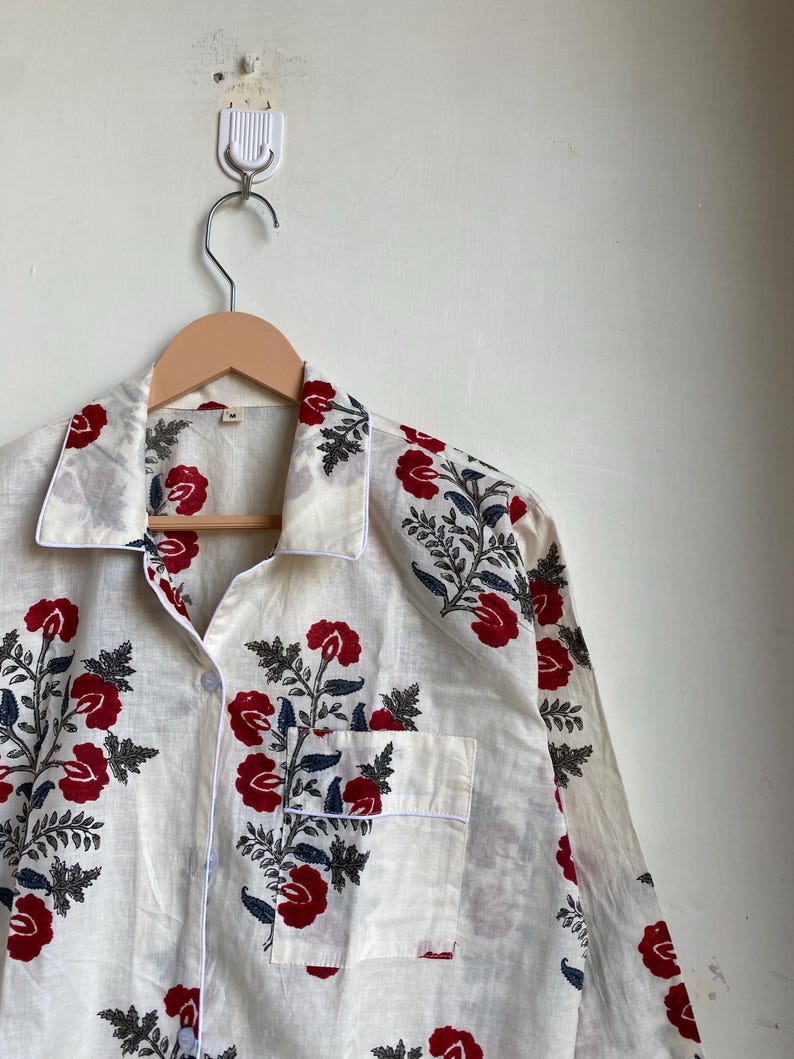 May include: Cream-colored pajama top with a red floral pattern and a white trim. The long-sleeved top has a button-down front, a chest pocket, and is hanging on a wooden hanger.