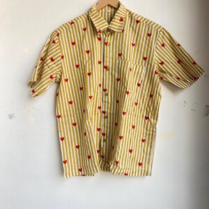 May include: A short-sleeved button-down shirt with a yellow and white striped pattern. Small red hearts are scattered across the fabric. The shirt has a single chest pocket and is hanging on a wooden hanger.
