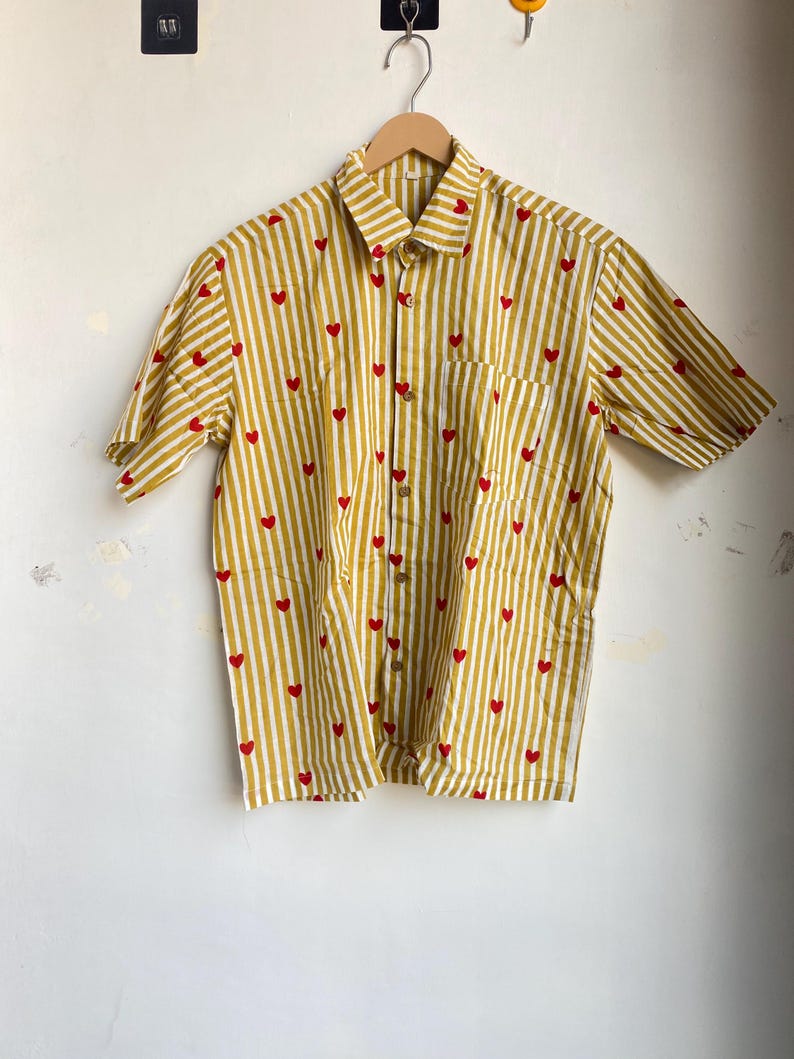 May include: A short-sleeved button-down shirt with a yellow and white striped pattern. Small red hearts are scattered across the fabric. The shirt has a single chest pocket and is hanging on a wooden hanger.