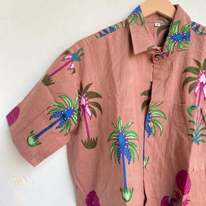 May include: A short-sleeved button-down shirt in a muted rose color, adorned with a repeating pattern of stylized palm trees in blue, green, and purple. The shirt has a classic collar and a single chest pocket. The shirt is hanging on a wooden hanger.