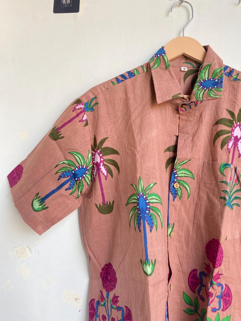 May include: A short-sleeved button-down shirt in a muted rose color, adorned with a repeating pattern of stylized palm trees in blue, green, and purple. The shirt has a classic collar and a single chest pocket. The shirt is hanging on a wooden hanger.