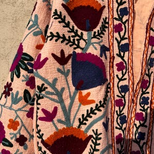May include: A pink embroidered jacket with a floral pattern in shades of blue, green, orange, and purple. The jacket has a long sleeve and a button-up front.