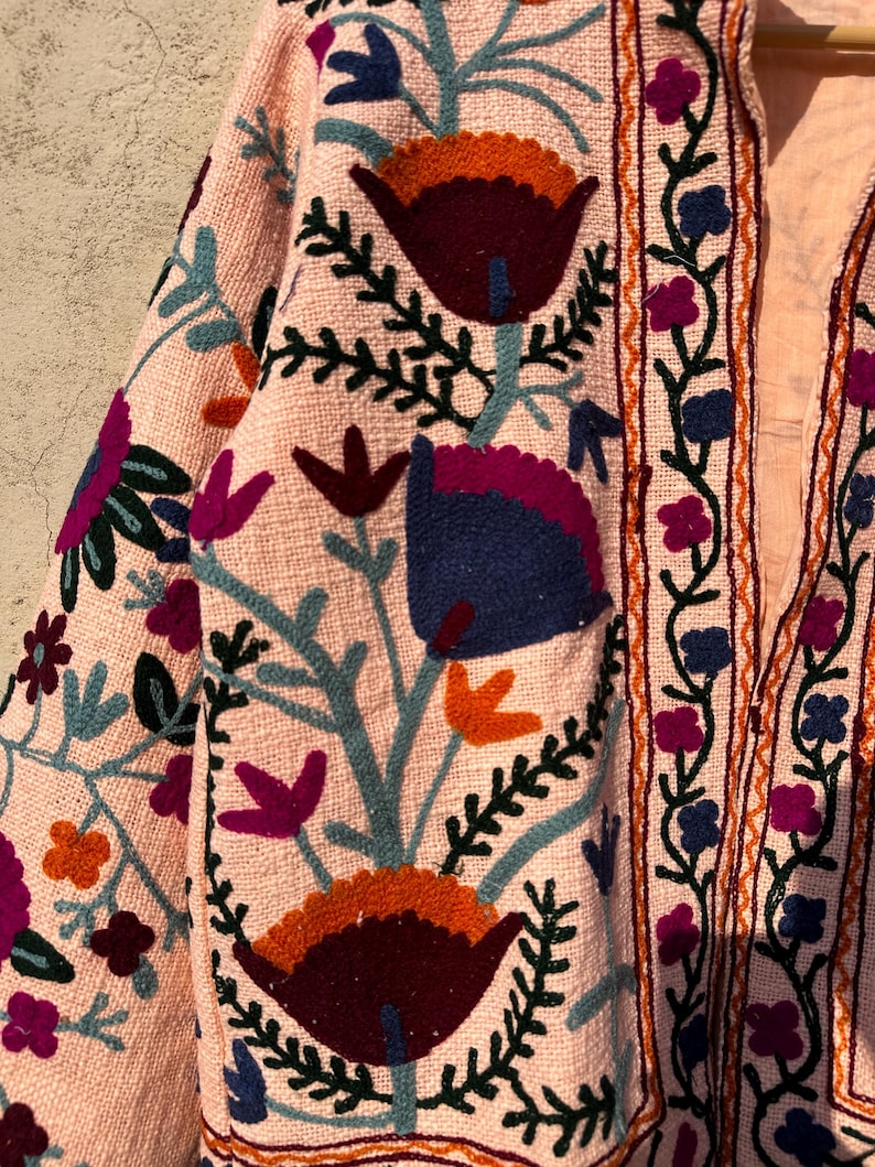 May include: A pink embroidered jacket with a floral pattern in shades of blue, green, orange, and purple. The jacket has a long sleeve and a button-up front.