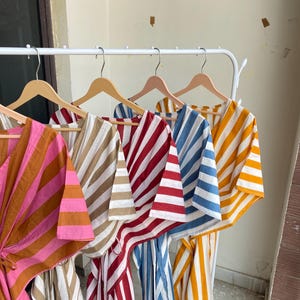 May include: A collection of striped caftans in various colors, including pink and brown, beige and white, red and white, blue and white, and yellow and white. The caftans are hanging on wooden hangers on a white rack.