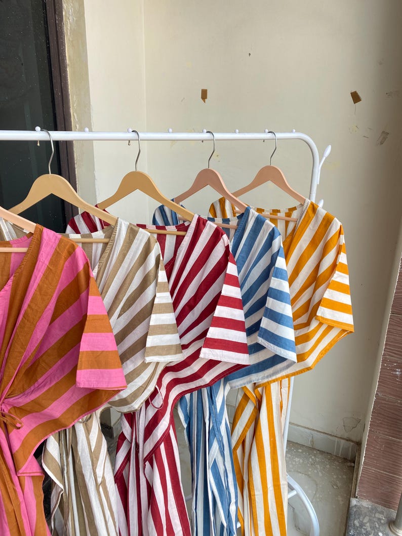 May include: A collection of striped caftans in various colors, including pink and brown, beige and white, red and white, blue and white, and yellow and white. The caftans are hanging on wooden hangers on a white rack.