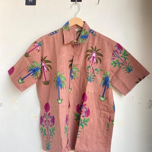 May include: A short-sleeved button-down shirt in a muted rose color, adorned with a repeating pattern of stylized palm trees and floral designs in shades of green, blue, and pink. The shirt has a classic collar and a single chest pocket.