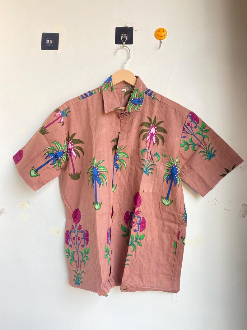 May include: A short-sleeved button-down shirt in a muted rose color, adorned with a repeating pattern of stylized palm trees and floral designs in shades of green, blue, and pink. The shirt has a classic collar and a single chest pocket.
