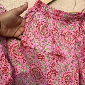 May include: Pink cotton pants with a floral print in shades of red and green. The pants have an elastic waistband.