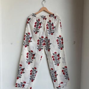May include: White pajama pants with a repeating floral pattern in red and navy blue. The pants are hanging on a wooden hanger against a white wall. The floral design is a stylized cluster of flowers and leaves.