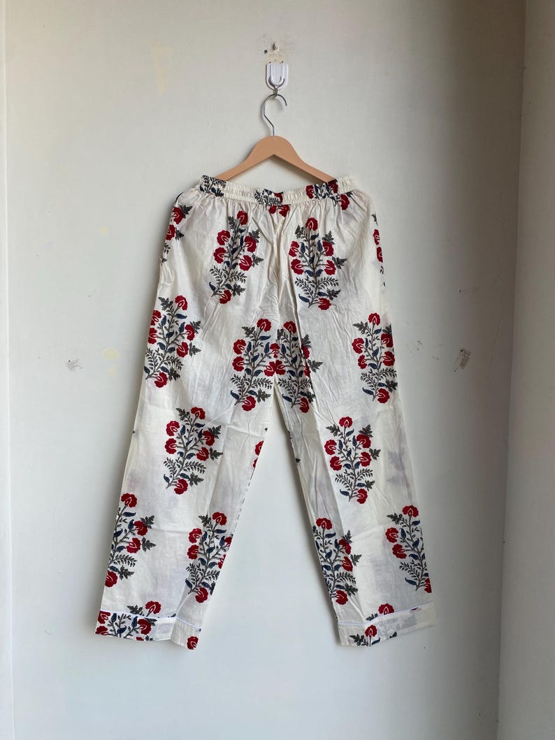 May include: White pajama pants with a repeating floral pattern in red and navy blue. The pants are hanging on a wooden hanger against a white wall. The floral design is a stylized cluster of flowers and leaves.