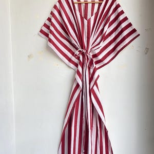 May include: A red and white striped kaftan dress hanging on a wooden hanger. The dress features a V-neck and a tie at the waist. The fabric has vertical stripes of red and white.