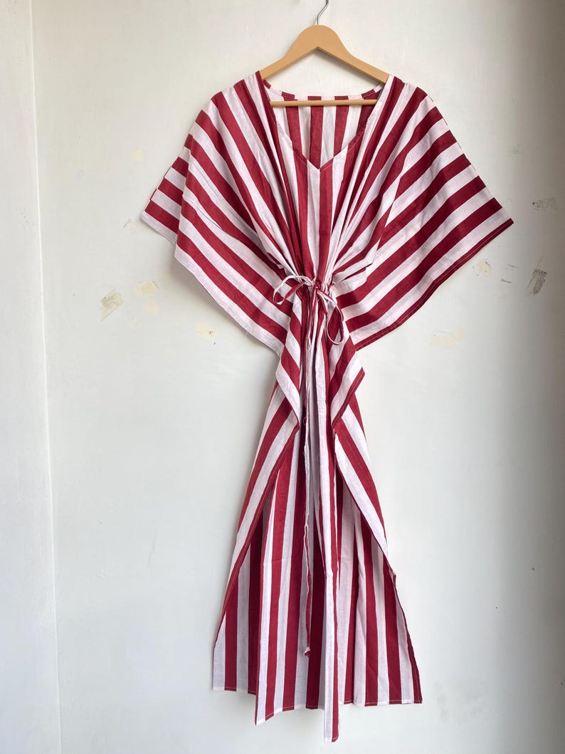 May include: A red and white striped kaftan dress hanging on a wooden hanger. The dress features a V-neck and a tie at the waist. The fabric has vertical stripes of red and white.