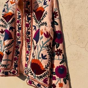 May include: A pink embroidered jacket with floral patterns in shades of blue, purple, orange, and green. The jacket has a mandarin collar and long sleeves.