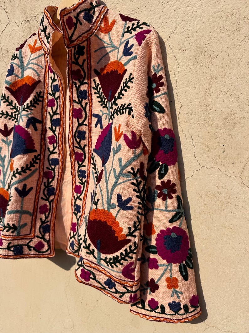 May include: A pink embroidered jacket with floral patterns in shades of blue, purple, orange, and green. The jacket has a mandarin collar and long sleeves.