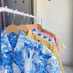 May include: A collection of button-down shirts in various patterns and colors hanging on a white rack. The shirts include a blue and white tropical print, yellow and white stripes, and floral designs. Each shirt is on a wooden hanger.