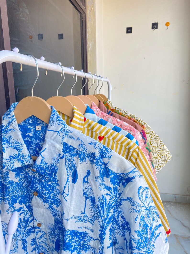 May include: A collection of button-down shirts in various patterns and colors hanging on a white rack. The shirts include a blue and white tropical print, yellow and white stripes, and floral designs. Each shirt is on a wooden hanger.