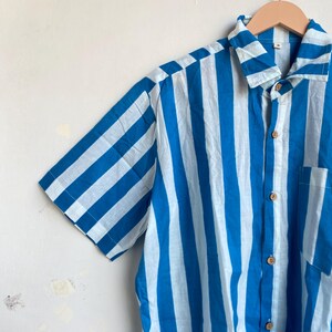 May include: Short-sleeved shirt with vertical stripes in light blue and white. The shirt has a button-down front, a collar, and a chest pocket. The shirt is hanging on a wooden hanger.