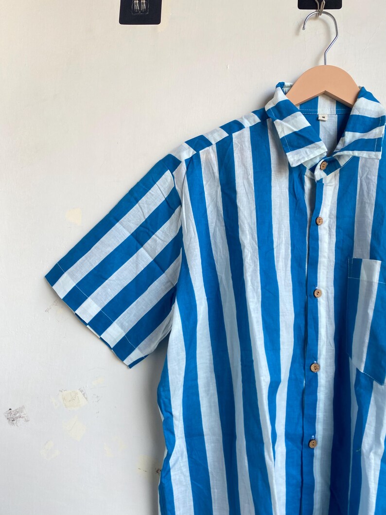 May include: Short-sleeved shirt with vertical stripes in light blue and white. The shirt has a button-down front, a collar, and a chest pocket. The shirt is hanging on a wooden hanger.