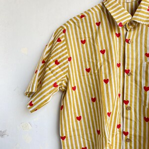 May include: Yellow and white striped button-up shirt with red heart accents. The short-sleeved shirt is hanging on a wooden hanger. The shirt has a collared neckline and wooden buttons.