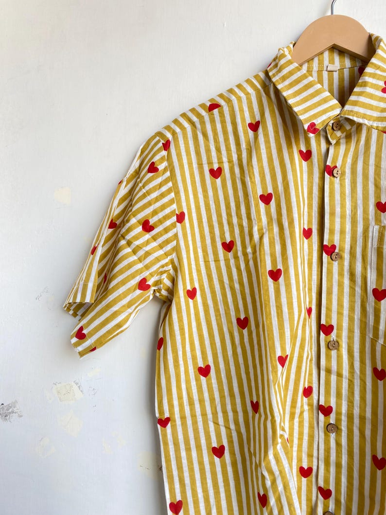 May include: Yellow and white striped button-up shirt with red heart accents. The short-sleeved shirt is hanging on a wooden hanger. The shirt has a collared neckline and wooden buttons.