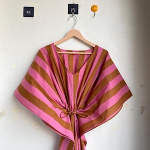 May include: A pink and brown striped caftan dress hanging on a wooden hanger. The dress has a V-neck and a tie at the waist. The stripes are horizontal across the body and sleeves. The background is a white wall.
