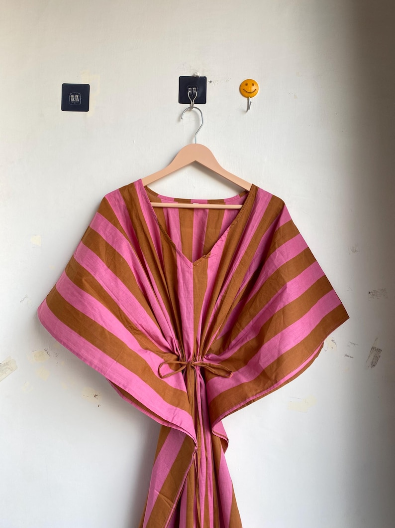 May include: A pink and brown striped caftan dress hanging on a wooden hanger. The dress has a V-neck and a tie at the waist. The stripes are horizontal across the body and sleeves. The background is a white wall.