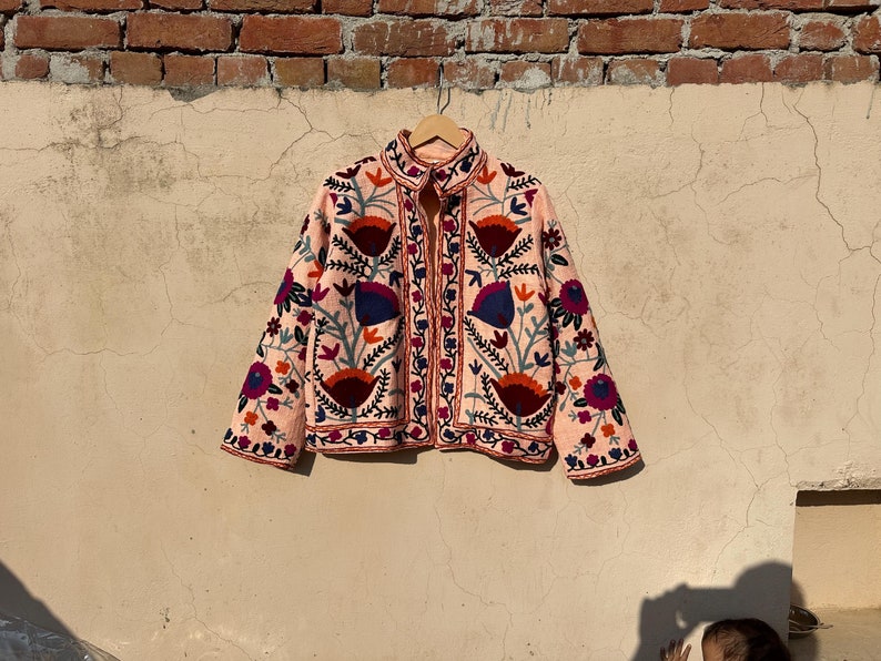May include: A pink embroidered jacket with floral patterns in shades of red, blue, and green. The jacket has a zipper closure and a stand-up collar.