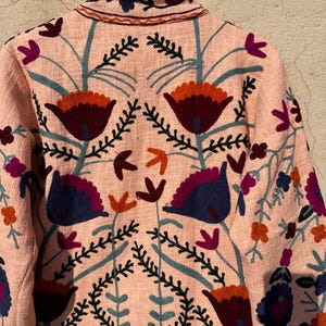 May include: A pink embroidered jacket with floral designs in shades of blue, red, orange, and green.