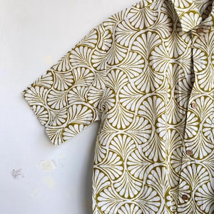 May include: Short-sleeved button-down shirt with a repeating fan-shaped pattern in olive green and white. The shirt has a classic collar and wooden buttons. The fabric appears to be lightweight and suitable for warm weather.