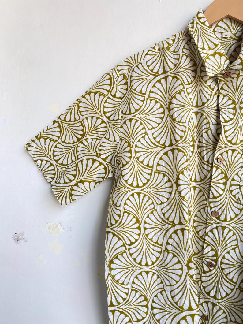 May include: Short-sleeved button-down shirt with a repeating fan-shaped pattern in olive green and white. The shirt has a classic collar and wooden buttons. The fabric appears to be lightweight and suitable for warm weather.