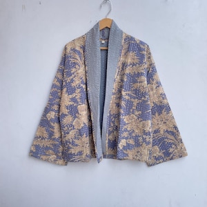 Peut inclure: Veste à motifs floraux dans les tons pervenche et beige, avec un col et une doublure gris. La veste est suspendue à un cintre en bois contre un mur blanc. Le motif floral est visible sur toute la surface de la veste.