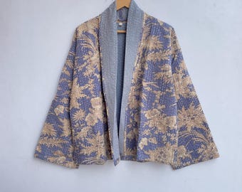 Pode incluir: Casaco com padrão floral em tons de azul-violeta e bege, com gola e forro cinzentos. O casaco está pendurado num cabide de madeira contra uma parede branca. O desenho floral é visível em toda a superfície do casaco.