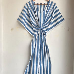 May include: A blue and white striped caftan dress hanging on a wooden hanger. The dress has a V-neck and a tie at the waist. The fabric is a light, airy material, perfect for warm weather. The stripes run vertically down the length of the dress.