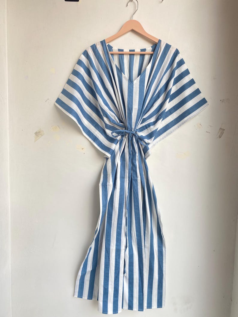 May include: A blue and white striped caftan dress hanging on a wooden hanger. The dress has a V-neck and a tie at the waist. The fabric is a light, airy material, perfect for warm weather. The stripes run vertically down the length of the dress.