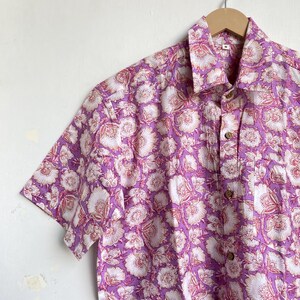 May include: A short-sleeved button-down shirt with a floral pattern. The shirt features a vibrant design of white and pink flowers on a purple background. It is hanging on a wooden hanger against a white wall.