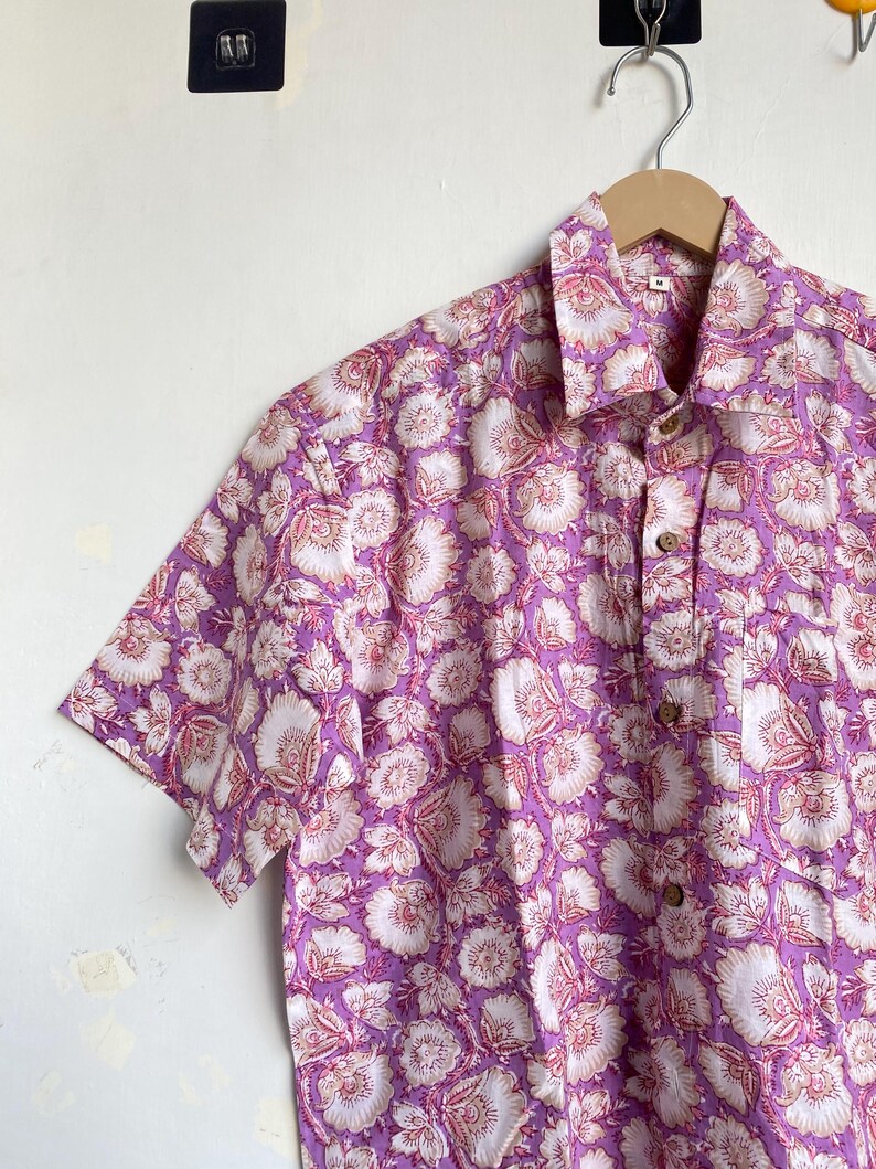 May include: A short-sleeved button-down shirt with a floral pattern. The shirt features a vibrant design of white and pink flowers on a purple background. It is hanging on a wooden hanger against a white wall.