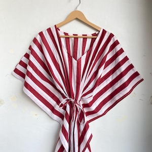 May include: A red and white striped kaftan dress with a V-neck and tie waist, hanging on a wooden hanger. The dress features wide, vertical stripes and a loose, flowing design, perfect for warm weather.