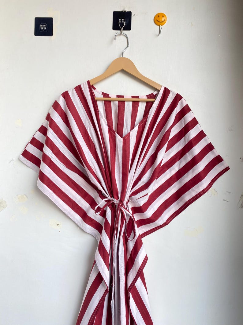 May include: A red and white striped kaftan dress with a V-neck and tie waist, hanging on a wooden hanger. The dress features wide, vertical stripes and a loose, flowing design, perfect for warm weather.