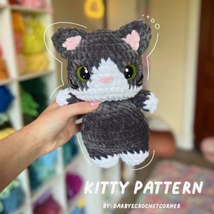 May include: A handmade crochet cat plushie in gray, white, and pink. The cat has large green eyes and is holding a white paw. The text "KITTY PATTERN" and "BY: DARBYSCROCHETCORNER" are visible. This soft toy is perfect for cat lovers.