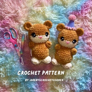 May include: Two crocheted brown hamster plushies with white feet and black eyes. One hamster is wearing a purple and white party hat. The plushies are on a rainbow coloured fluffy surface. CROCHET PATTERN BY: DARBYSCROCHETCORNER