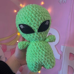 Peut inclure: Une peluche extraterrestre crochetée vert citron avec de grands yeux noirs et des accents de paillettes argentées. L'extraterrestre a de petits bras et un corps arrondi. Le fond est un mur rose avec des lumières décoratives et des autocollants en forme de lapin.