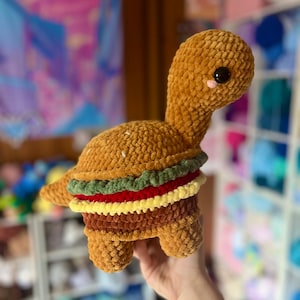 Op de afbeelding: Een gehaakte knuffel schildpad in de vorm van een hamburger. De schildpad is bruin met groene sla, rode tomaat, gele kaas en bruine vleesvulling.