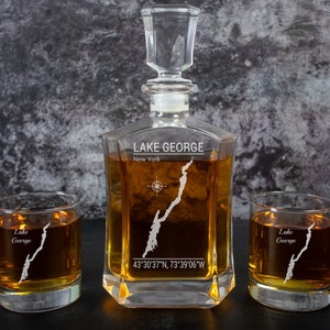 May include: A crystal decanter and two matching glasses with an etched design of Lake George, New York. The decanter and glasses are filled with amber liquid. The text "LAKE GEORGE" and "New York" are etched on the decanter. The coordinates "43'30'37"N, 73°39'06"W" are also etched on the decanter.