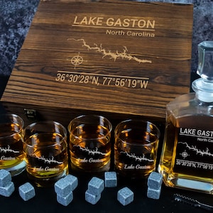 May include: A wooden box set with a decanter, four glasses, and whiskey stones. The set is engraved with "Lake Gaston, North Carolina" and coordinates. The glasses and decanter are filled with amber liquid.