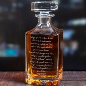 May include: A clear glass decanter with a square base and a crystal stopper. The decanter is filled with a golden liquid and features engraved text. The text includes a date, and a message. The decanter is likely a personalized gift.