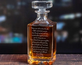 May include: A clear glass decanter with a square base and a crystal stopper. The decanter is filled with a golden liquid and features engraved text. The text includes a date, and a message. The decanter is likely a personalized gift.