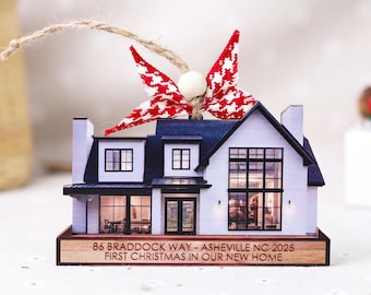May include: A white and black house ornament with a navy blue roof and a red and white patterned bow. The ornament has the text "86 BRADDOCK WAY - ASHEVILLE NC 2025 FIRST CHRISTMAS IN OUR NEW HOME" on the base.