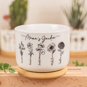 May include: White ceramic plant pot with the text "Mama's Garden" and floral illustrations. The pot has names below each flower. It sits on a wooden coaster and is surrounded by plants.